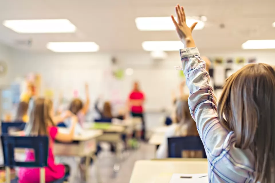 View from back of classroom of young children at desks raising hands.